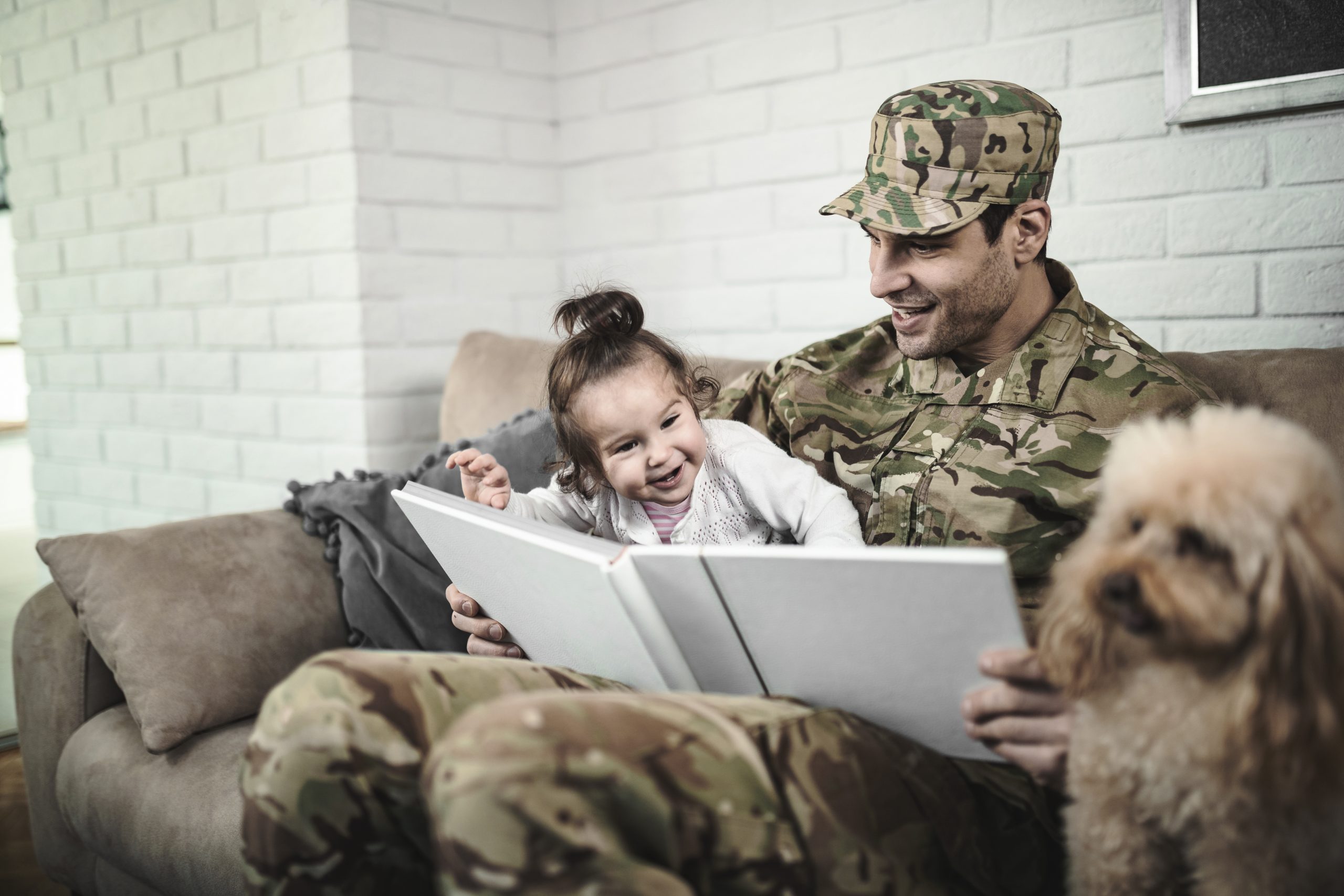 Young happy soldier having fun with his small daughter while looking at pictures in photo album at home.
